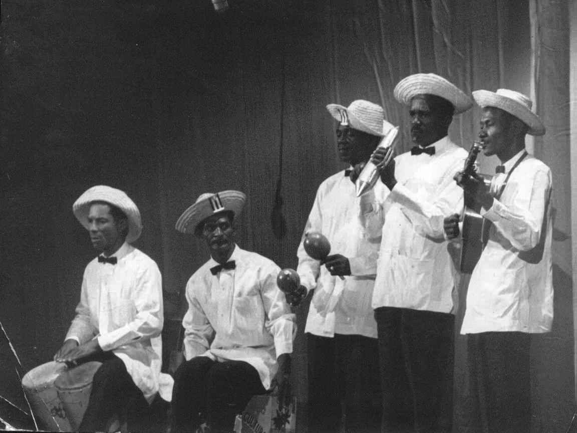 A photograph of five musicians performing in white guayabera shirts, dark pants, and straw hats